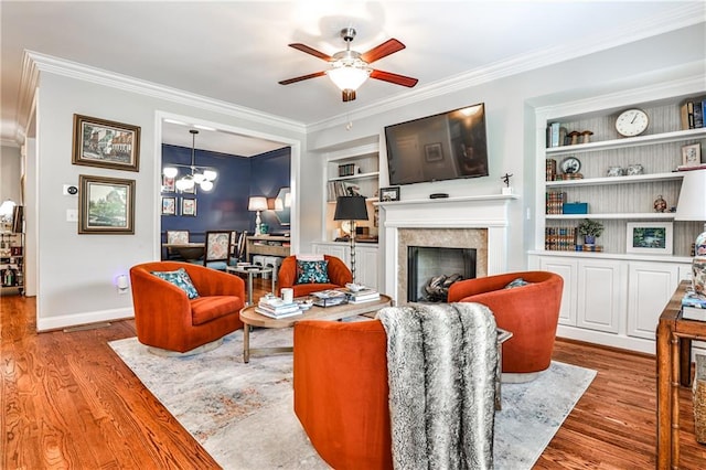 living area with built in features, wood finished floors, ceiling fan with notable chandelier, a glass covered fireplace, and ornamental molding