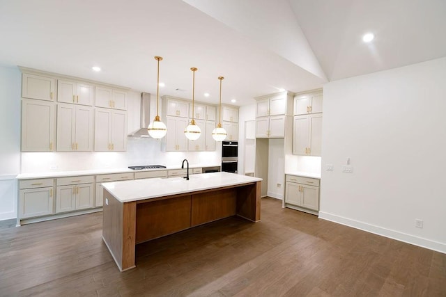 kitchen with wall chimney exhaust hood, appliances with stainless steel finishes, a kitchen island with sink, light countertops, and pendant lighting