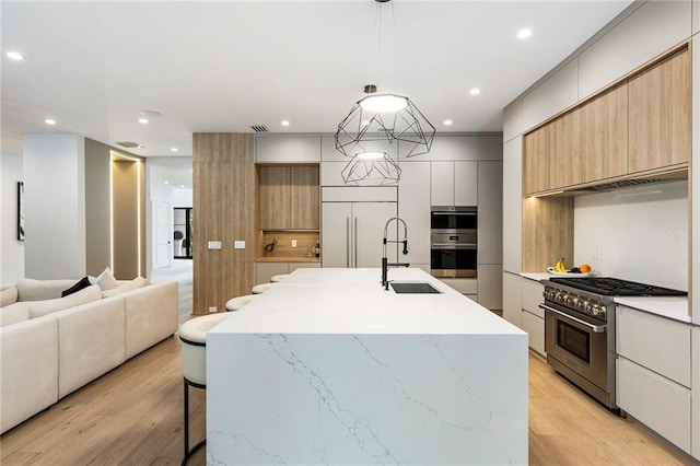 kitchen featuring high end appliances, hanging light fixtures, sink, light wood-type flooring, and an island with sink