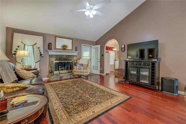 living room with a stone fireplace, a ceiling fan, wood finished floors, baseboards, and high vaulted ceiling