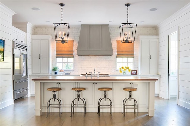 kitchen featuring custom range hood, stainless steel built in fridge, a center island with sink, a chandelier, and hanging light fixtures