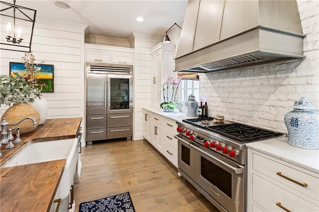 kitchen with high quality appliances, custom exhaust hood, butcher block counters, a chandelier, and tasteful backsplash