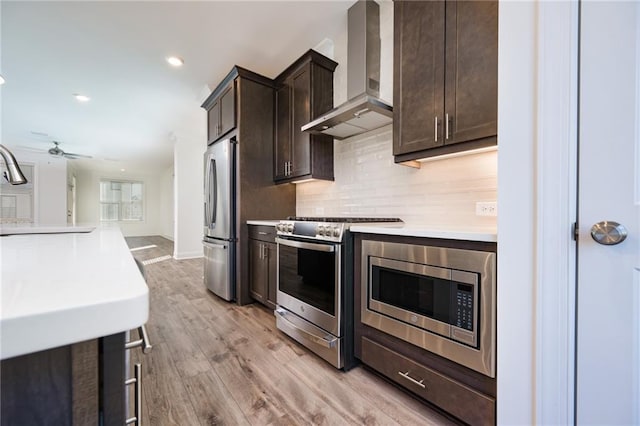 kitchen with dark brown cabinetry, light wood-type flooring, appliances with stainless steel finishes, decorative backsplash, and wall chimney range hood