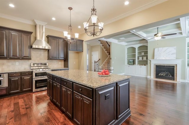 kitchen featuring a kitchen island, appliances with stainless steel finishes, decorative light fixtures, dark brown cabinets, and wall chimney exhaust hood