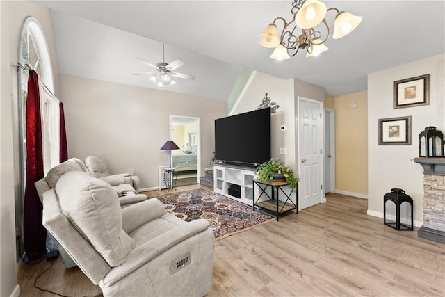 living room with light wood-type flooring, ceiling fan with notable chandelier, and a stone fireplace