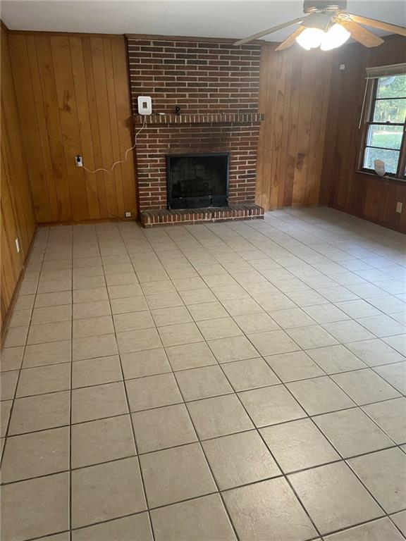 unfurnished living room with wooden walls, a brick fireplace, light tile patterned flooring, brick wall, and ceiling fan