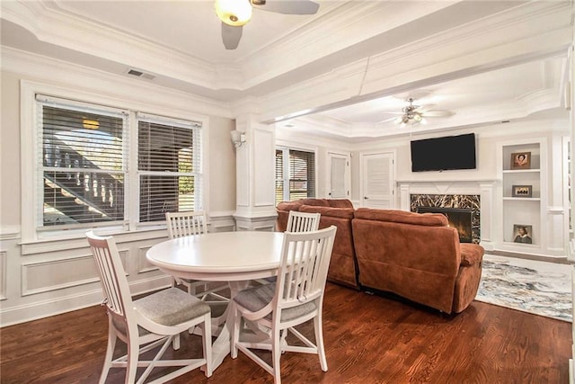 dining space featuring dark wood-type flooring, built in features, a premium fireplace, ceiling fan, and a raised ceiling