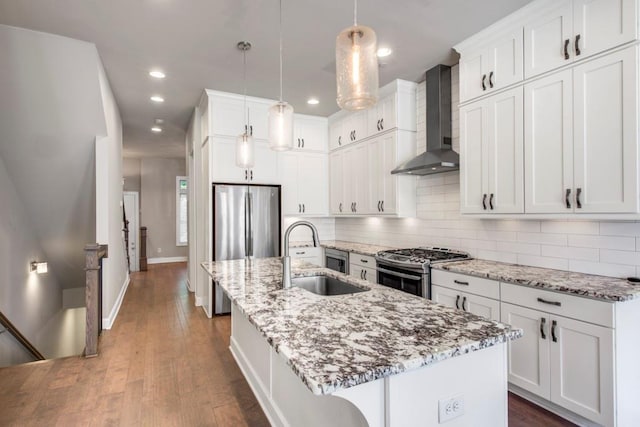 kitchen with stainless steel appliances, sink, wall chimney range hood, hanging light fixtures, and an island with sink