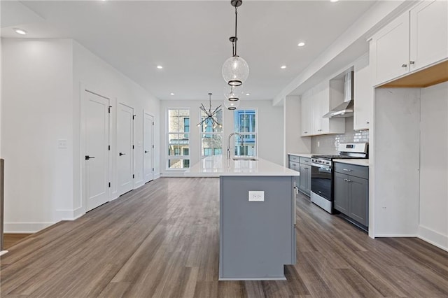 kitchen with stainless steel stove, wall chimney exhaust hood, an island with sink, decorative light fixtures, and sink