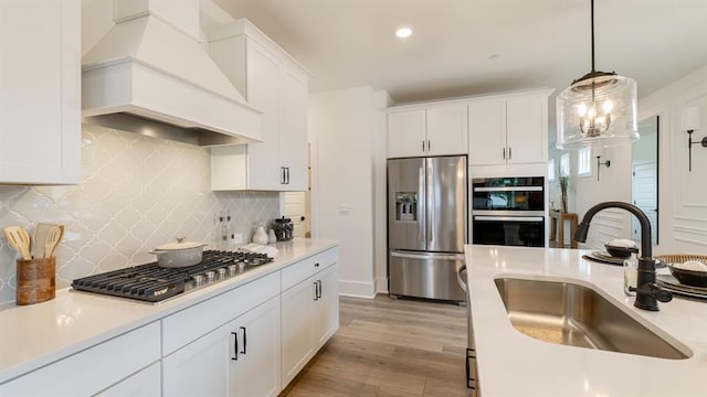 kitchen featuring stainless steel appliances, a sink, custom range hood, white cabinets, and decorative light fixtures