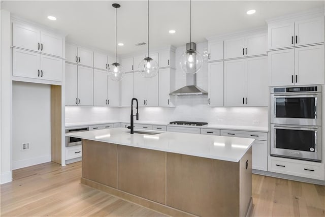 kitchen featuring stainless steel double oven, an island with sink, a sink, light countertops, and light wood-style floors