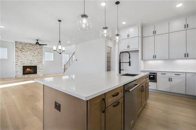 kitchen featuring a ceiling fan, light wood finished floors, a sink, a stone fireplace, and stainless steel dishwasher