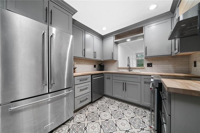 kitchen featuring appliances with stainless steel finishes, butcher block counters, gray cabinets, a sink, and wall chimney exhaust hood