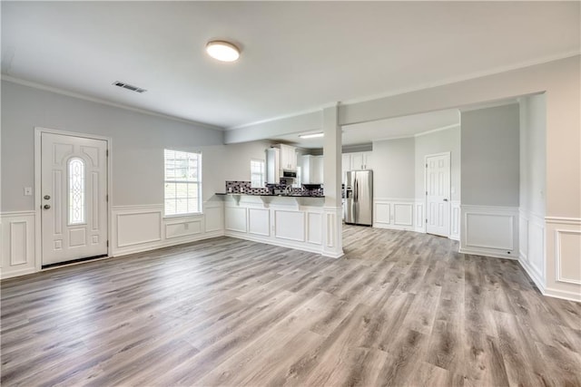 unfurnished living room with light wood-type flooring, visible vents, a wainscoted wall, and ornamental molding