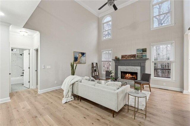 living area featuring light wood finished floors, ornamental molding, a ceiling fan, a stone fireplace, and baseboards