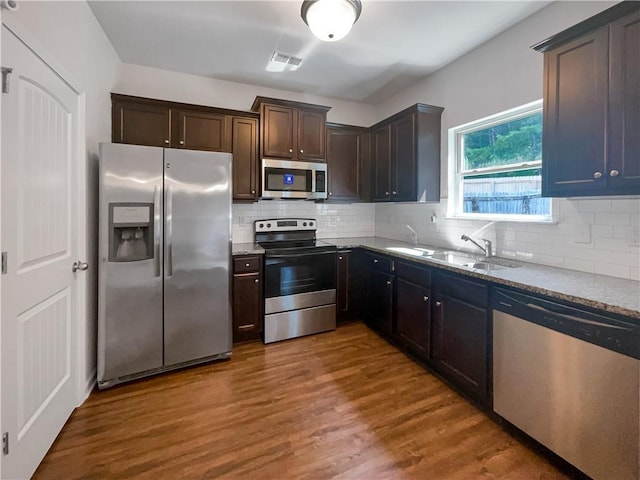 kitchen with appliances with stainless steel finishes, hardwood / wood-style floors, backsplash, light stone counters, and dark brown cabinetry