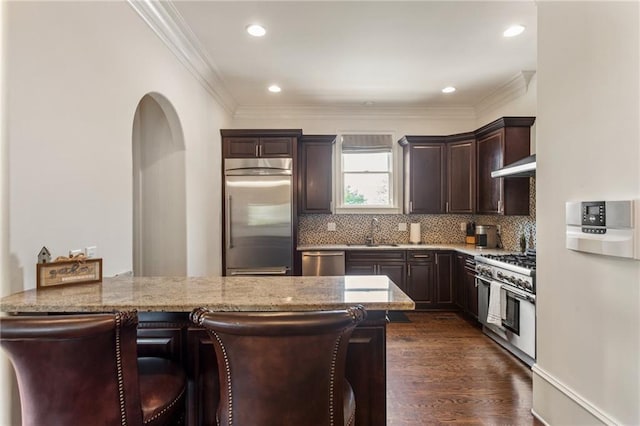 kitchen featuring high end appliances, dark brown cabinetry, range hood, light stone counters, and a sink
