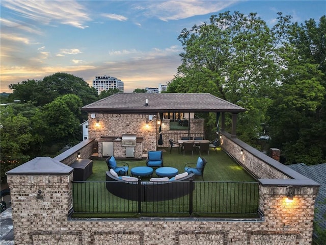 patio terrace at dusk featuring a yard, an outdoor kitchen, fence, and a grill