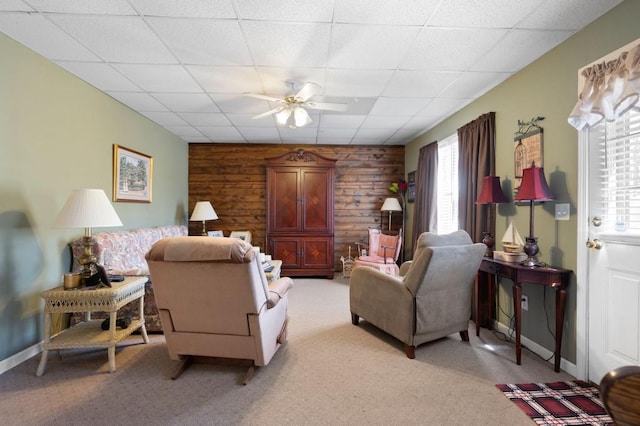 living room with a drop ceiling, wooden walls, light colored carpet, and a ceiling fan