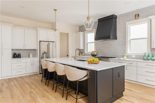 kitchen with premium range hood, a kitchen island with sink, white cabinetry, and a sink