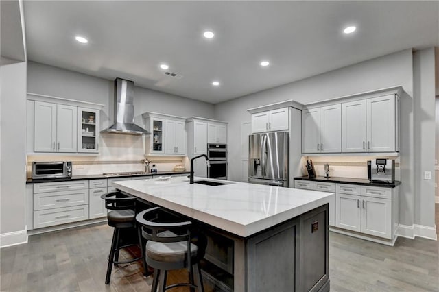 kitchen with wall chimney exhaust hood, an island with sink, appliances with stainless steel finishes, and glass insert cabinets