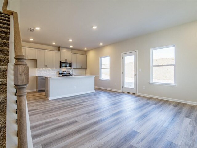 kitchen with gray cabinetry, decorative backsplash, stove, a kitchen island with sink, and light wood-type flooring