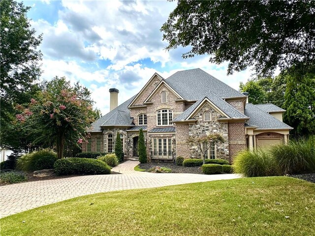 french country inspired facade featuring a garage, stone siding, a chimney, decorative driveway, and a front yard