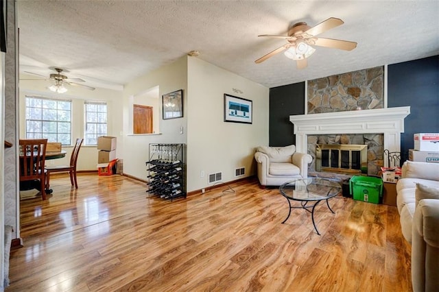 living room featuring ceiling fan, a stone fireplace, a textured ceiling, and light wood-type flooring