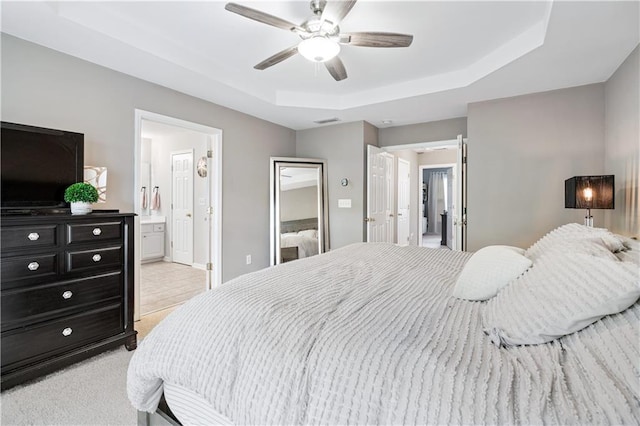 bedroom with ceiling fan, light colored carpet, visible vents, a tray ceiling, and ensuite bath