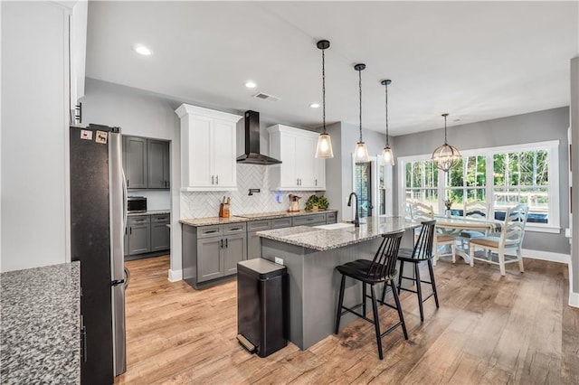 kitchen featuring a breakfast bar area, light wood-type flooring, stainless steel refrigerator, a kitchen island with sink, and wall chimney range hood