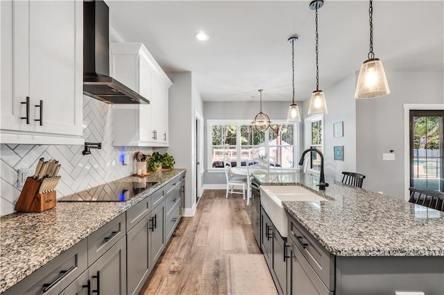 kitchen with wall chimney exhaust hood, decorative light fixtures, black electric stovetop, a kitchen island with sink, and backsplash