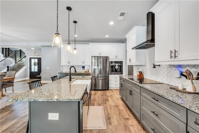 kitchen featuring a kitchen island with sink, hanging light fixtures, stainless steel appliances, tasteful backsplash, and wall chimney exhaust hood