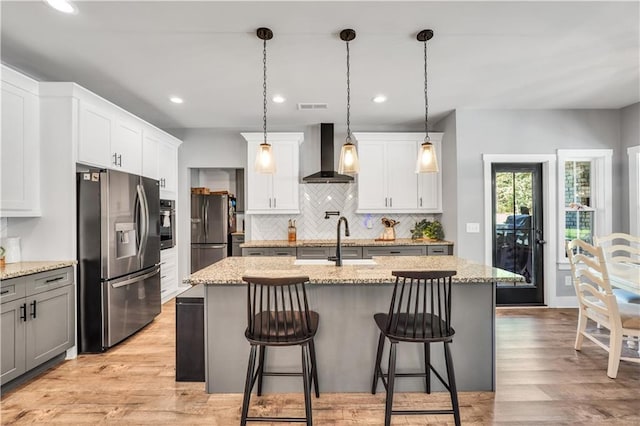 kitchen with hanging light fixtures, appliances with stainless steel finishes, a kitchen island with sink, and wall chimney range hood