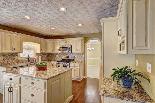 kitchen featuring arched walkways, appliances with stainless steel finishes, a kitchen island, a sink, and light stone countertops