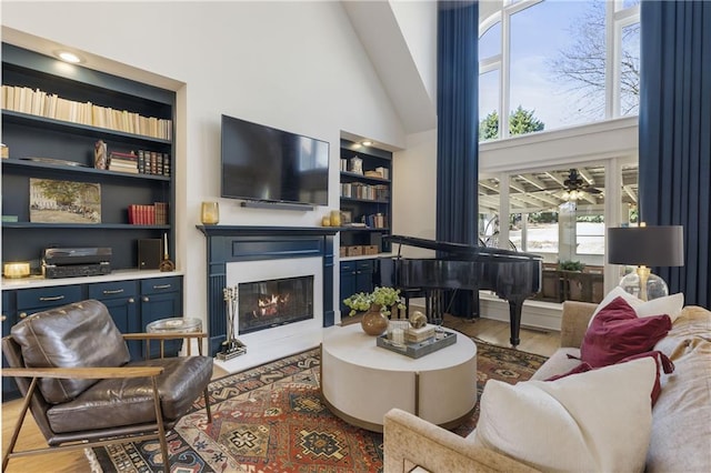 living room featuring built in features, a ceiling fan, wood finished floors, high vaulted ceiling, and a glass covered fireplace