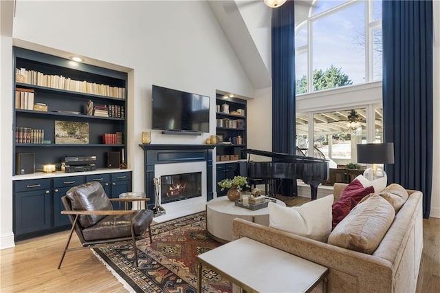 living room featuring built in shelves, high vaulted ceiling, light wood-style flooring, a glass covered fireplace, and ceiling fan