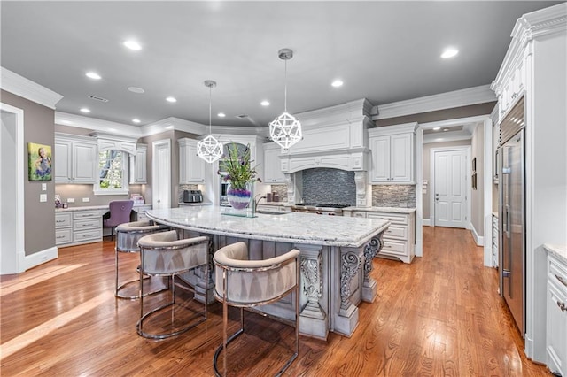 kitchen with light wood-style flooring, premium range hood, backsplash, stainless steel built in refrigerator, and a center island with sink