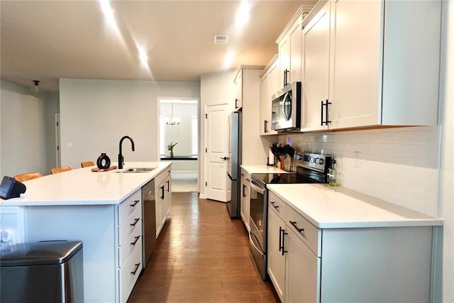 kitchen with a sink, backsplash, dark wood finished floors, appliances with stainless steel finishes, and a kitchen island with sink