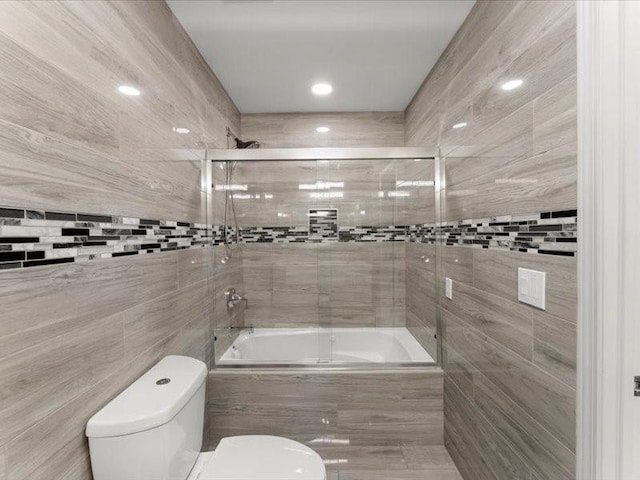 bathroom with toilet, bath / shower combo with glass door, and tile walls