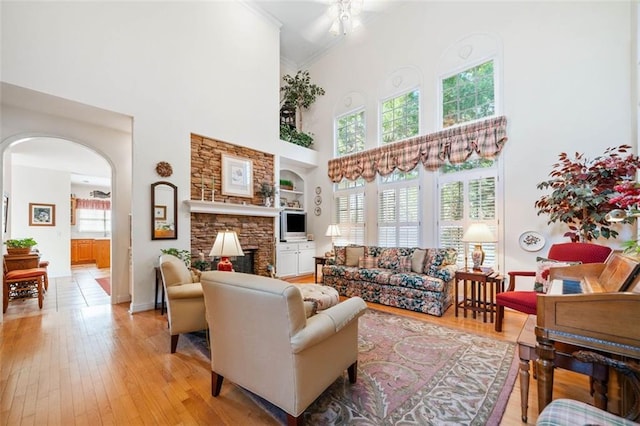 living room featuring a stone fireplace, a towering ceiling, built in features, ornamental molding, and light hardwood / wood-style flooring