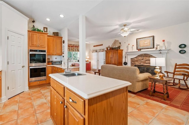 kitchen featuring a stone fireplace, an island with sink, light tile patterned flooring, double oven, and sink