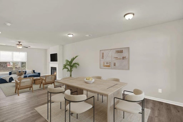 dining room featuring a glass covered fireplace, ceiling fan, baseboards, and wood finished floors