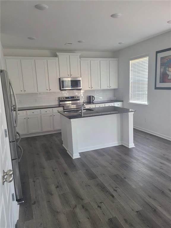 kitchen with a center island with sink, backsplash, dark countertops, dark wood-style floors, and stainless steel appliances