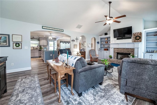 living room with a ceiling fan, arched walkways, lofted ceiling, a brick fireplace, and dark wood-type flooring