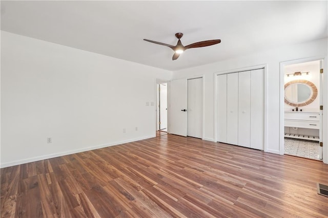 unfurnished bedroom featuring two closets, wood-type flooring, ensuite bath, and ceiling fan