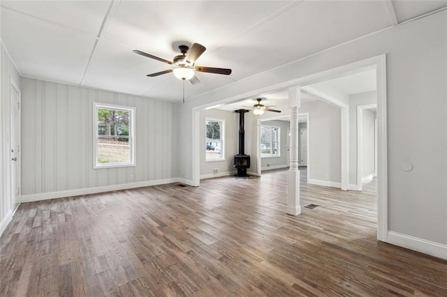 spare room featuring baseboards, a ceiling fan, wood finished floors, a wood stove, and ornate columns