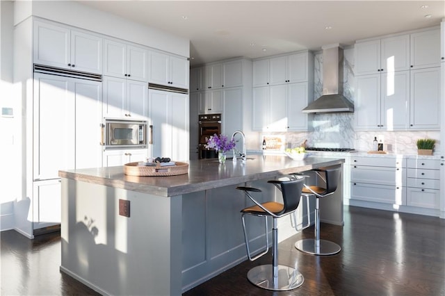 kitchen with white cabinetry, built in appliances, a large island with sink, and wall chimney exhaust hood