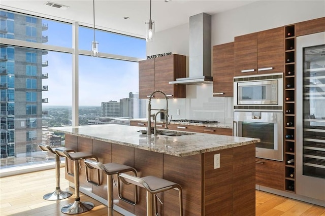 kitchen with a kitchen island with sink, light wood-type flooring, decorative light fixtures, stainless steel appliances, and wall chimney range hood