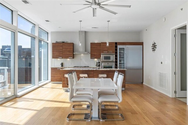 kitchen featuring pendant lighting, built in appliances, ceiling fan, wall chimney exhaust hood, and light hardwood / wood-style floors