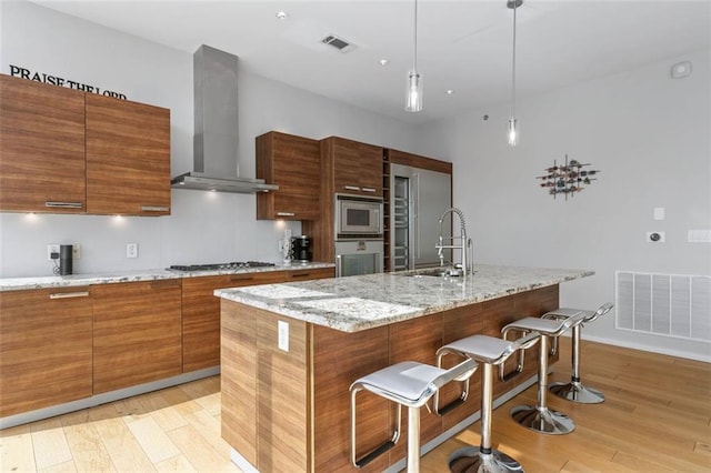 kitchen featuring light hardwood / wood-style flooring, decorative light fixtures, stainless steel appliances, wall chimney exhaust hood, and a center island with sink